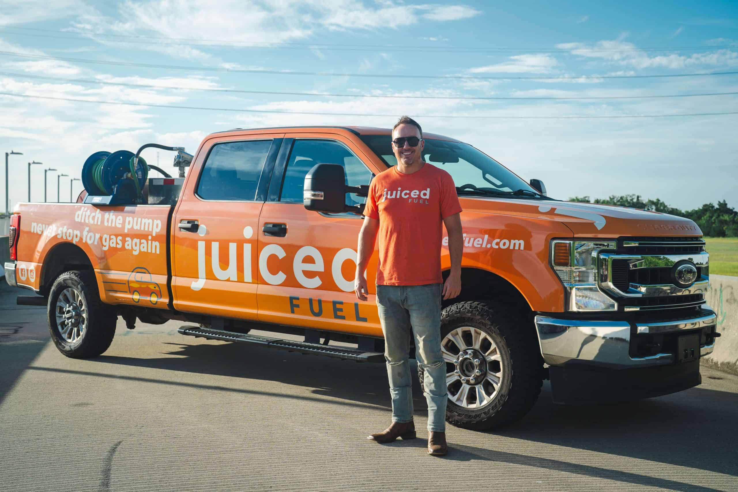 Korey McDavid standing in front of a Juiced Fuel delivery truck on a sunny day, wearing a branded orange t-shirt and sunglasses.